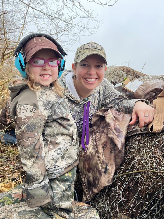 A child and a woman pose in hunting clothes outdoors.