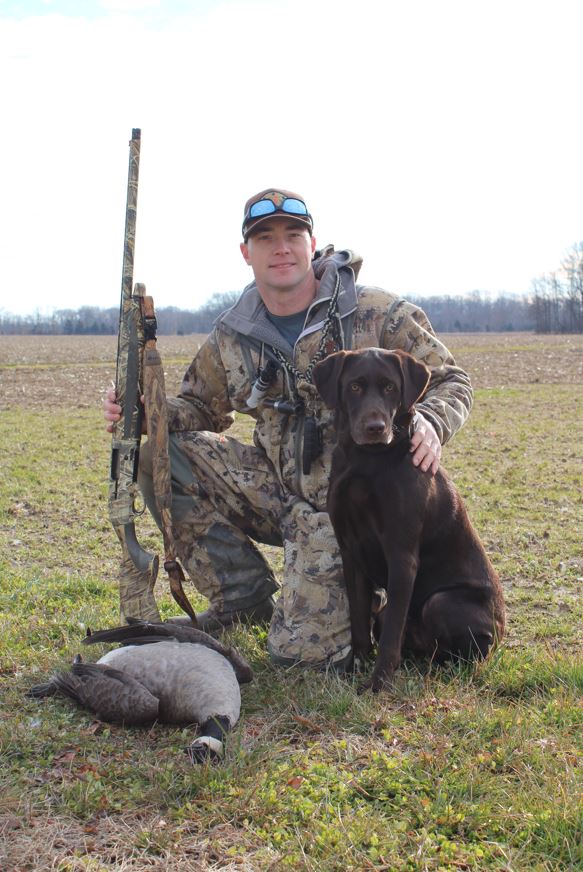 A man kneels in a field with a hunting rifle, a dog and a dead goose.