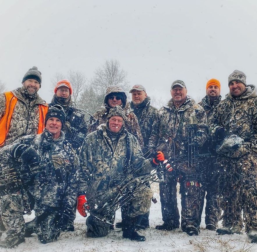 A group of men in archery hunting gear pose in a snow-covered field in a snow storm.