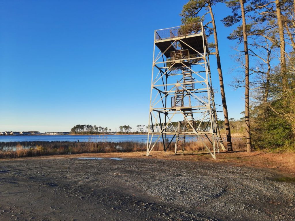 A metal tower with stairs and a deck on top stands by open water in a coastal landscape.