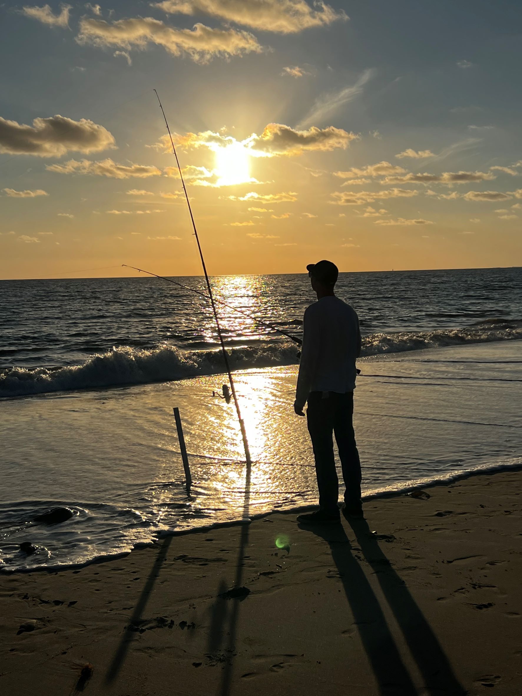 A person stands with a fishing rod at the shore, silhouetted by the sun. 