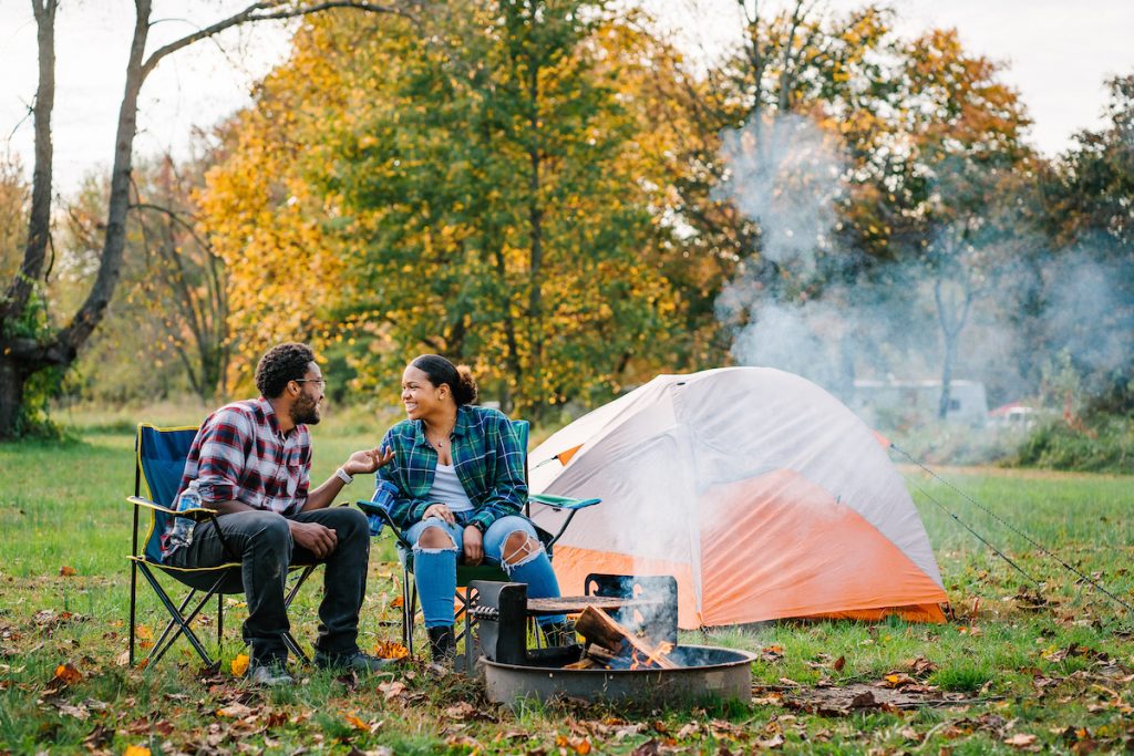 A man and a woman sit in folding chairs by a campfire with a small tent next to them and trees showing fall foliage behind them.